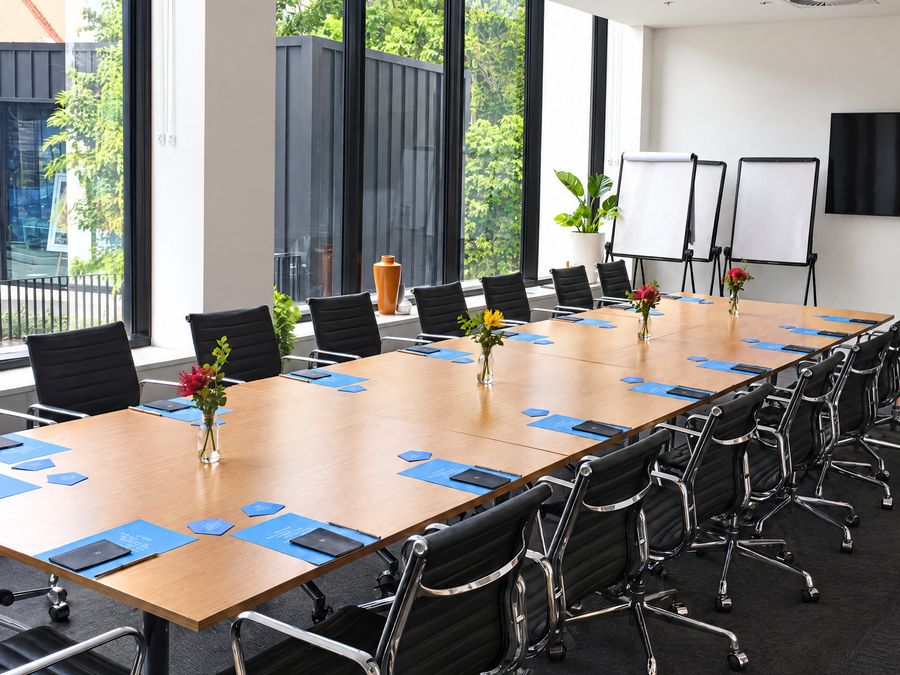Boardroom table set with notepads and flowers beside floor to ceiling windows, flip charts at the far end