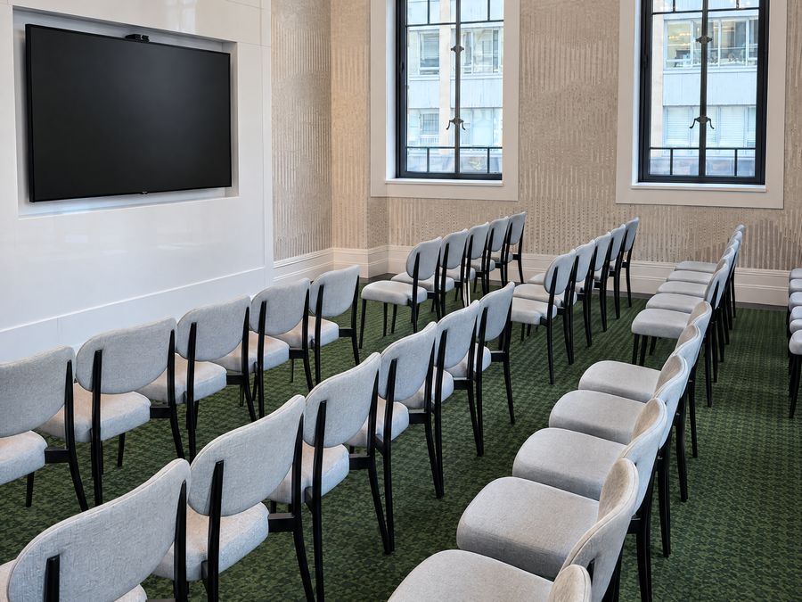 Conference room set theatre style, rows of upholstered chairs facing a wall screen, with tall windows