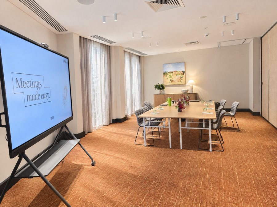 Adina Sydney Darling Harbour, meeting room set with a long table, an interactive display on a mobile stand in the foreground and full length curtains