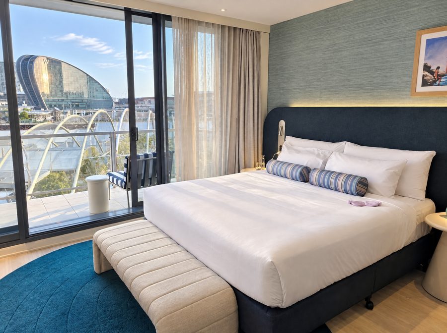 Adina Sydney Darling Harbour, guest room with a king bed and a navy headboard, sliding doors onto a balcony looking across to the curved glass tower of W Sydney