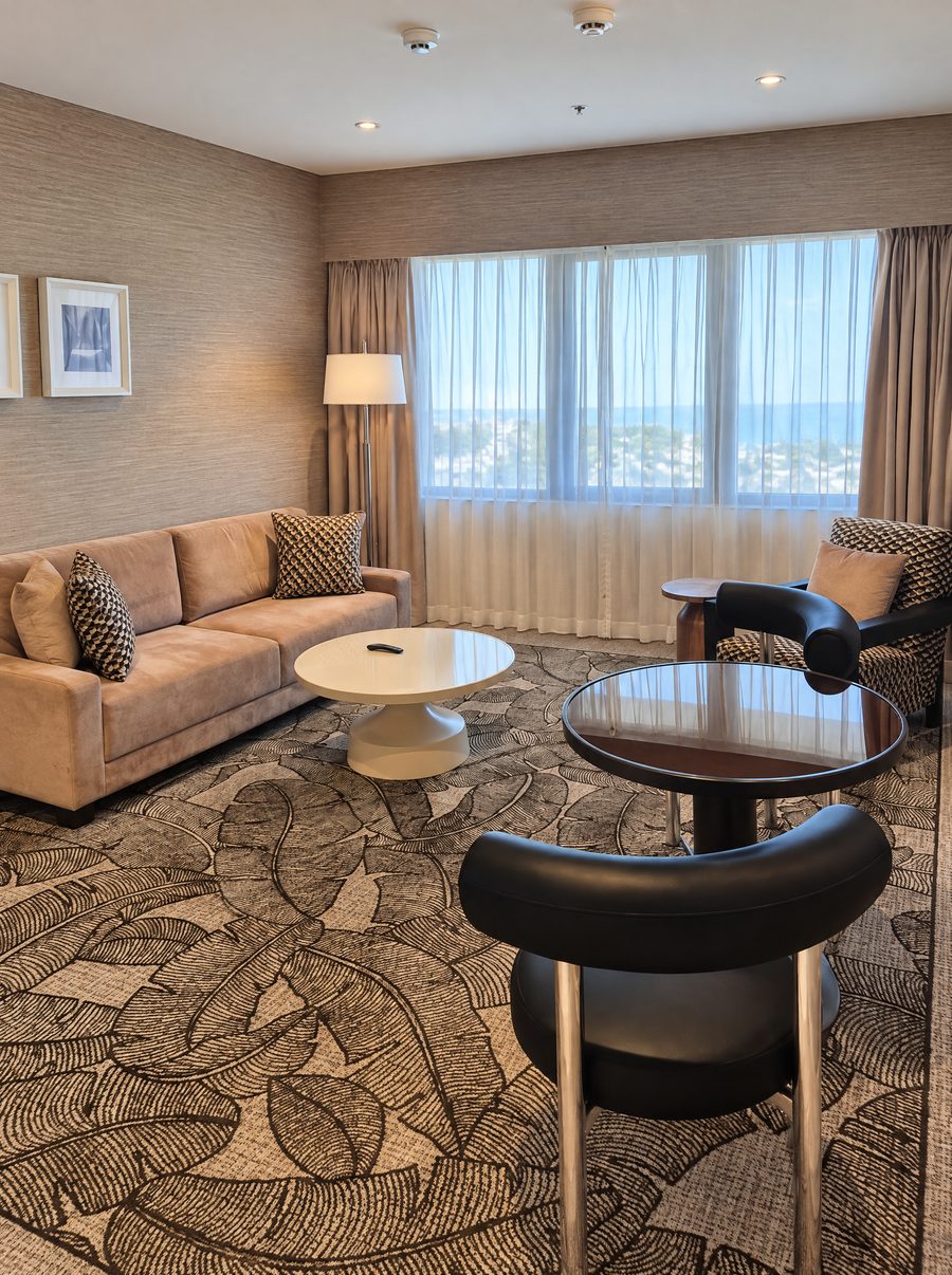 DoubleTree by Hilton Esplanade Darwin, suite living room with a suede sofa, a round glass table and sheer curtains over a sea view