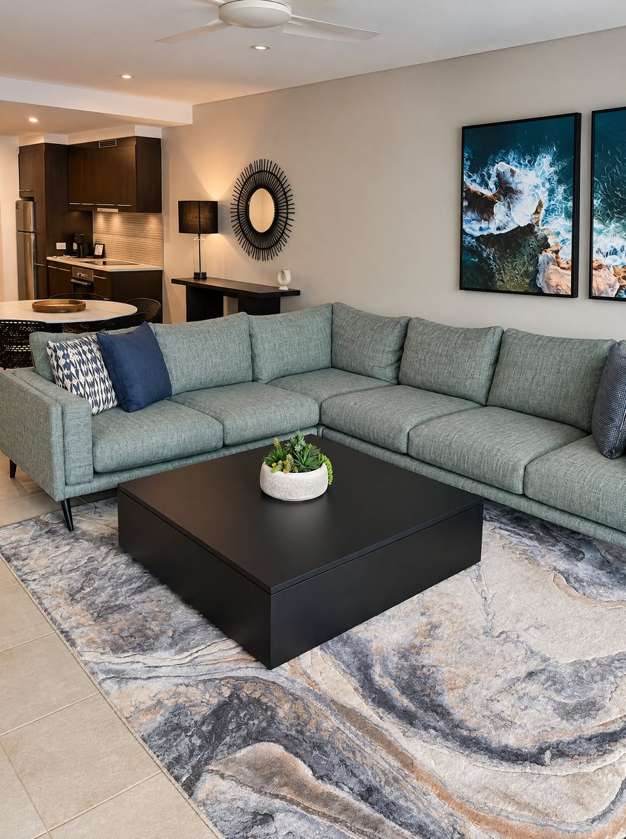 Pullman Port Douglas Sea Temple Resort & Spa, green corner sofa and black coffee table on a marbled rug, two framed aerial ocean prints on the wall and the kitchen beyond
