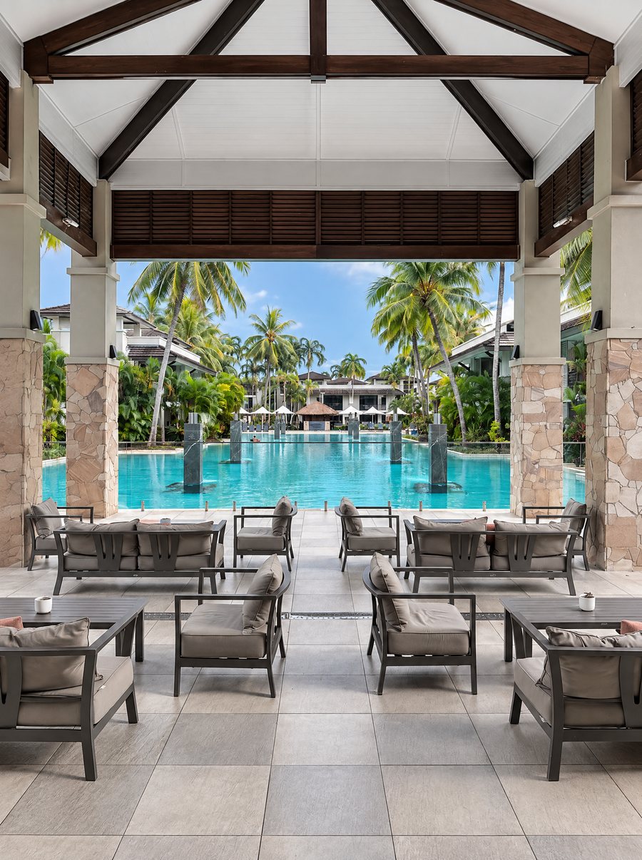 Pullman Port Douglas Sea Temple Resort & Spa, covered open-air lounge of outdoor sofas under a timber-beamed roof, looking over the lagoon pool and palms