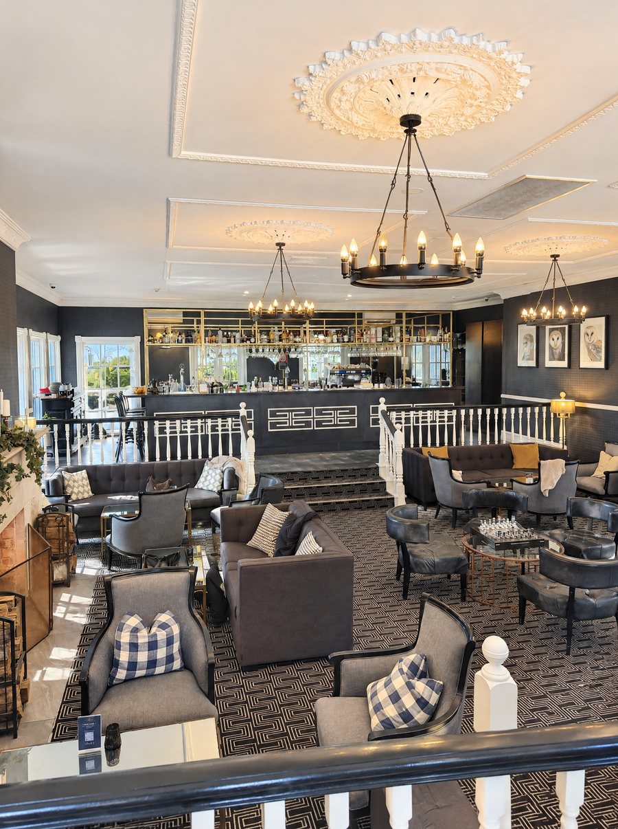 voco Kirkton Park Hunter Valley, lounge bar with a black and gold back bar on a raised level, grey armchairs and sofas with check cushions on a geometric carpet, a wrought iron chandelier overhead