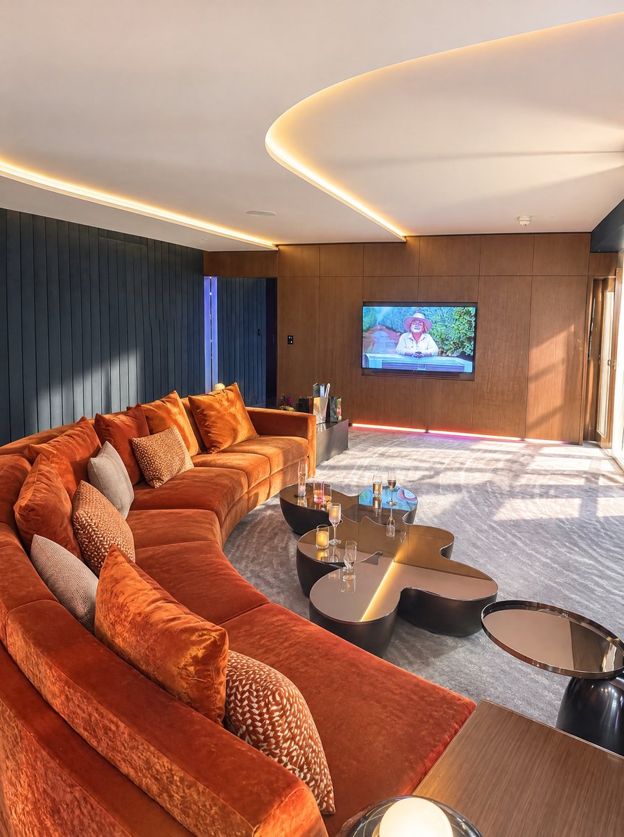 W Sydney, suite living room with a long curved rust velvet sofa, a sculptural bronze coffee table and a lit curved ceiling, a screen set into a timber panelled wall