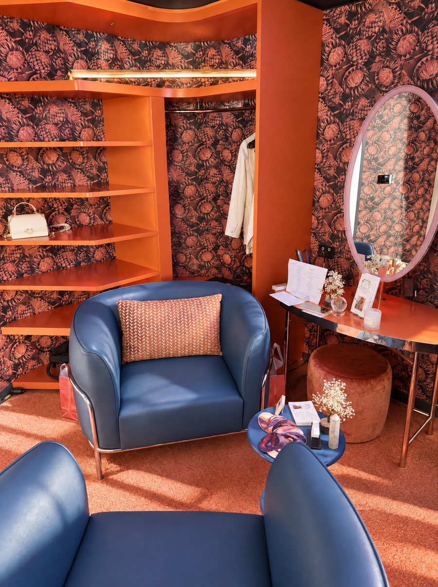 W Sydney, dressing area with orange lacquered shelving and a hanging rail against botanical print wallpaper, a blue leather armchair, a copper vanity and an oval mirror